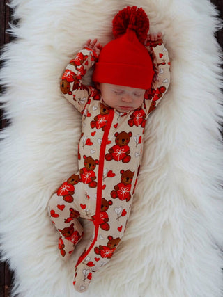 A baby sleeps peacefully on a fluffy white surface, wearing SpearmintLOVEs Basic 2-Way Zip Footie - Valentine Bear, crafted from soft bamboo viscose in beige and red. The outfit features hearts, flowers, and bears. With arms up and legs spread comfortably, the scene is adorable.