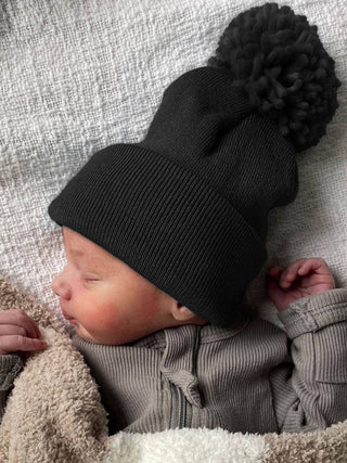 A newborn baby sleeps peacefully on a soft, textured blanket, wearing a cozy gray onesie and SpearmintLOVEs Bamboo Babys First Hat with a black pom-pom. The babys hands are gently curled near their face.