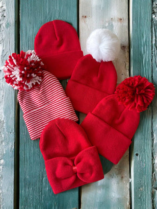 On a rustic wooden surface, Babys First Hat by SpearmintLOVE showcases festive red winter hats in various styles, including one with a pom-pom, a bow, and red and white stripes. Crafted from soft, stretchy fabric for ultimate comfort.