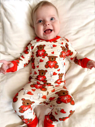 A cheerful baby in SpearmintLOVEs cream Long Sleeve 2-Piece Set - Valentine Bear with red cuffs is lying on a white blanket, smiling widely and capturing the joy of family matching.