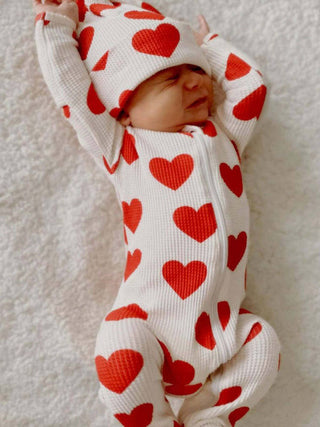 A baby wearing a SpearmintLOVE Organic Waffle 2-Way Zip Footie - Red Heart lies peacefully on a soft, white textured surface, eyes closed and arms above its head.