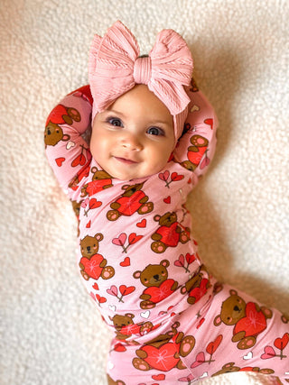 A happy baby, wearing the SpearmintLOVE Long Sleeve 2-Piece Set - Valentine Bear Pink, adorned with teddy bears and hearts, lies on a soft fabric. The pink headband with a large bow enhances the outfits charm, as her tiny arms rest gently above her head.