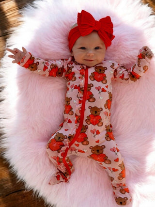 A baby smiles while lying on a fluffy pink blanket, wearing SpearmintLOVEs 2-Way Ruffle Zip Footie in Valentine Bear Pink. The onesie features a red headband, heart and bear patterns, buttery soft fabric, and a convenient 2-way zipper for easy changes.