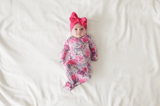 A baby in a Posh Peanut® Bamboo Basic Knotted Gown - Ellery (Floral) with pink, purple, and white flowers lies on a white blanket gazing upward. The gown allows easy diaper changes, complemented by a pink headband featuring a large bow.